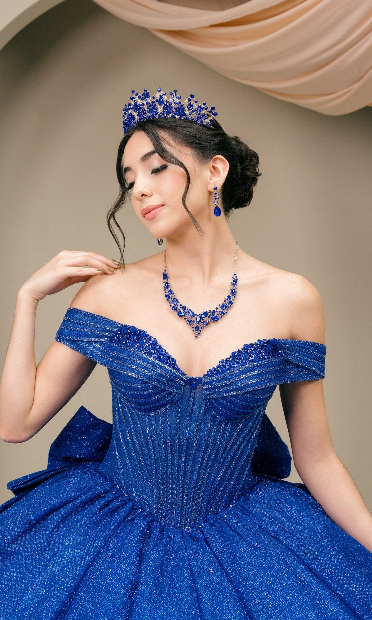 A woman poses with eyes closed in the Quinceanera Dress 8460JQ By Cinderella Couture, an off-the-shoulder blue ball gown styled with a glitter finish and matching jeweled tiara, earrings, and necklace against a neutral background.